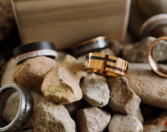 May include: A collection of rings in various styles and materials, displayed on a bed of small rocks. Rings include a gold band with black and red inlays, a black band with a wood-like inlay, and a silver ring with black accents.