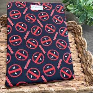 May include: A rectangular fabric pouch featuring a black background with a repeating red and white Deadpool logo pattern. The design includes the word Deadpool printed in red text throughout the fabric. A small white rectangular label is attached.