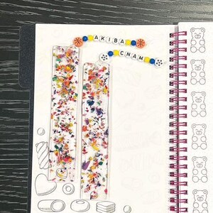 May include: Two clear resin bookmarks filled with colorful confetti sit on a white notebook page. The word "AKIBA CNAM" is spelled out with colorful beads. The notebook has a spiral binding and a pattern of bear illustrations.
