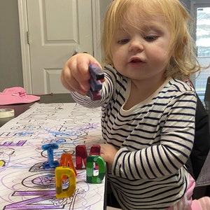May include: A young child in a striped top and pink trousers holds a crayon. The word "DAD" is spelled out in colourful wax crayons on a colouring sheet. The child is looking at the crayons.