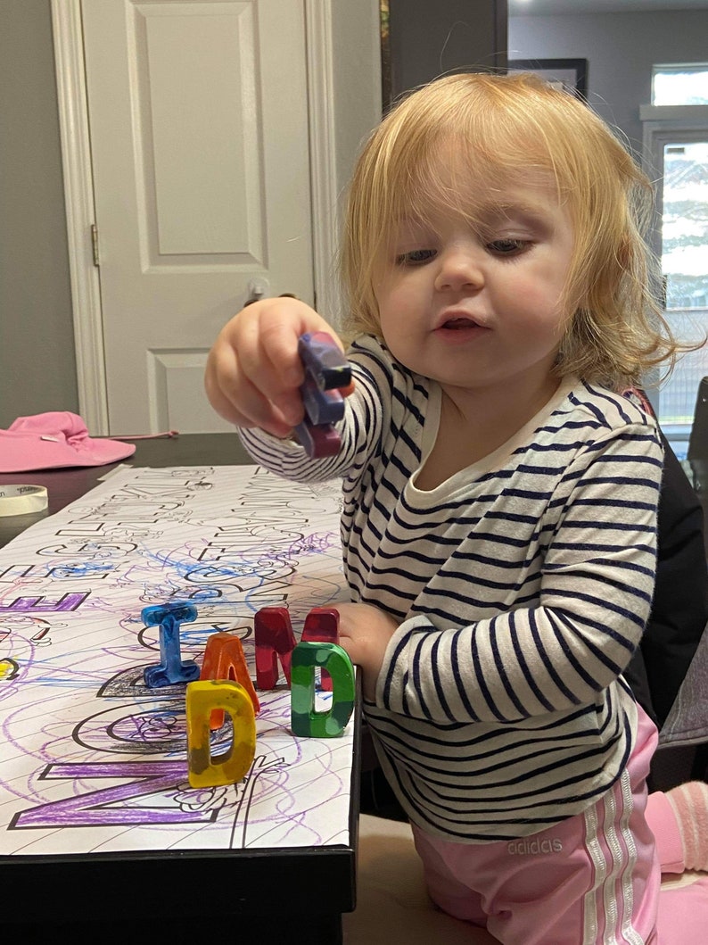 May include: A young child in a striped top and pink trousers holds a crayon. The word "DAD" is spelled out in colourful wax crayons on a colouring sheet. The child is looking at the crayons.
