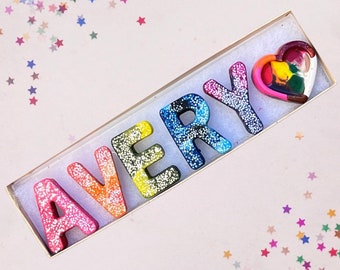 May include: A white frame with a rainbow glitter name "AVERY" spelled out in colorful letters. A small heart-shaped piece of rainbow glitter is in the upper right corner of the frame.