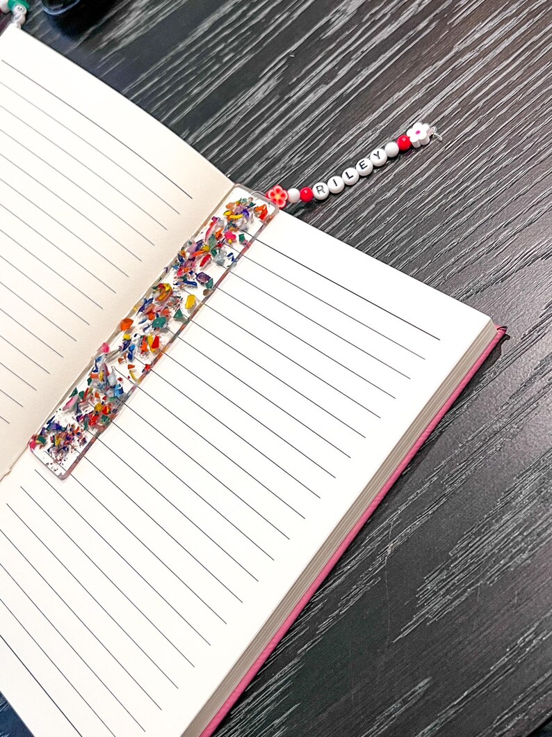 May include: A handmade bookmark with a clear resin body filled with colorful confetti. The bookmark is inserted into a lined notebook. A beaded string with the name "RILEY" and flower accents is attached.