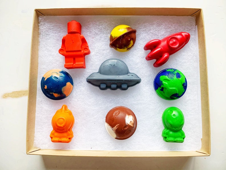 May include: A set of nine space-themed crayons in a brown box. The crayons are shaped like planets, a rocket, an alien, a spaceship, and a lego figure.