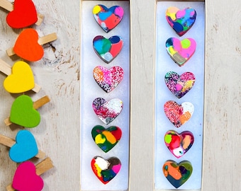 May include: Two rows of colorful heart-shaped wax melts, displayed in a white frame on a light wood background. The wax melts are arranged vertically in each row, with a variety of colors and patterns.