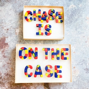May include: Two boxes of colourful crayons spelling out "CHASE IS ON THE CASE". The crayons are in the shape of capital letters and are a mix of red, yellow, and blue colours. The crayons are in a cardboard box with white paper.