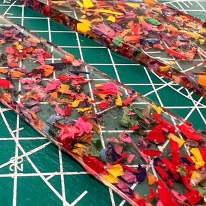 May include: Two clear, rectangular resin pieces filled with colorful crayon shavings. The shavings are a mix of red, pink, yellow, green, and purple. The pieces are on a green cutting mat with white grid lines.