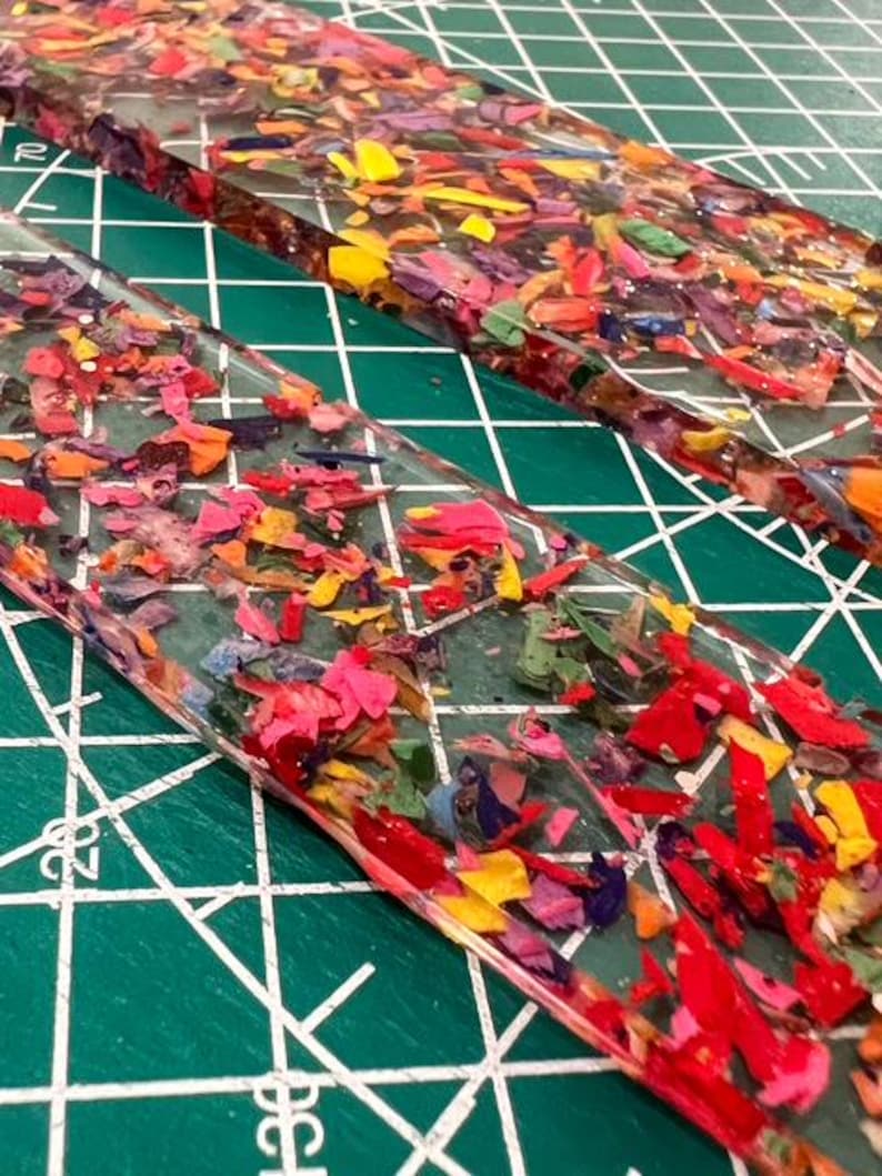May include: Two clear, rectangular resin pieces filled with colorful crayon shavings. The shavings are a mix of red, pink, yellow, green, and purple. The pieces are on a green cutting mat with white grid lines.