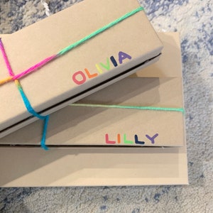 May include: Two rectangular, light brown gift boxes are stacked. The top box is tied with a rainbow-coloured string and has the name "OLIVIA" printed in rainbow colours. The bottom box has the name "LILLY" printed in rainbow colours.