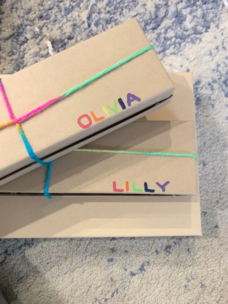 May include: Two rectangular, light brown gift boxes are stacked. The top box is tied with a rainbow-coloured string and has the name "OLIVIA" printed in rainbow colours. The bottom box has the name "LILLY" printed in rainbow colours.