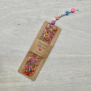 May include: A handmade bookmark with colorful confetti embedded in clear resin. The bookmark is attached to a string of beads spelling "joy" and flower-shaped beads. A brown paper tag reads "Handmade with love" and "Thank you."