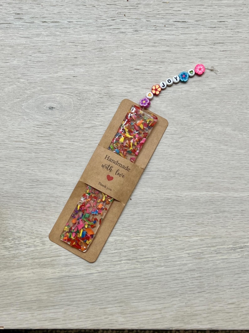 May include: A handmade bookmark with colorful confetti embedded in clear resin. The bookmark is attached to a string of beads spelling "joy" and flower-shaped beads. A brown paper tag reads "Handmade with love" and "Thank you."