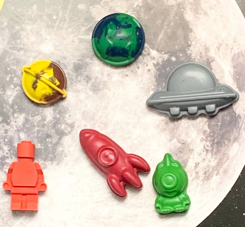 May include: A set of six space-themed crayons, including a rocket, a spaceship, an astronaut, and three planets. The crayons are all different colors and have a variety of designs.