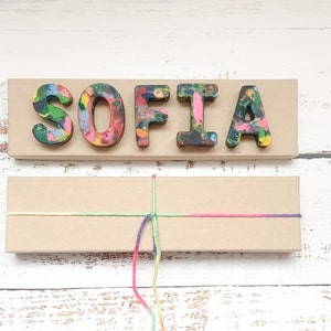 May include: A wooden sign displaying the name "SOFIA" in vibrant crayon letters. Beneath, a rectangular box is tied with a multi-coloured string, possibly a gift or craft item. The background is a white wooden surface.