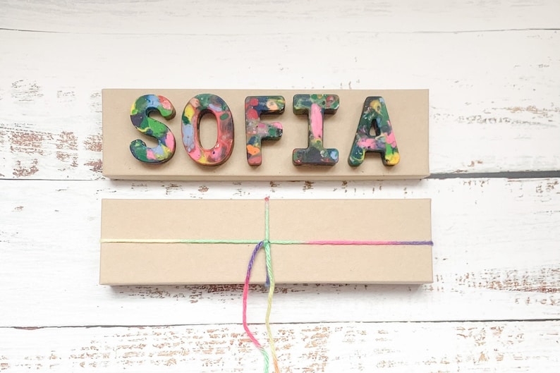 May include: A wooden sign displaying the name "SOFIA" in vibrant crayon letters. Beneath, a rectangular box is tied with a multi-coloured string, possibly a gift or craft item. The background is a white wooden surface.