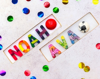 May include: A white background with colorful confetti and two names spelled out with colorful letters. The names are NOAH and AVA. A red ball and a soccer ball are placed above the names.