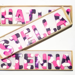 May include: Three boxes containing crayon names: "HAYLEY", "STELLA", and "SIENNA". The letter-shaped crayons have a marbled effect in pink, purple, and white. Each name is presented in its own box, offering a colourful and personalised gift.