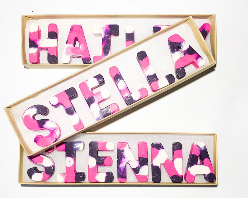 May include: Three boxes containing crayon names: "HAYLEY", "STELLA", and "SIENNA". The letter-shaped crayons have a marbled effect in pink, purple, and white. Each name is presented in its own box, offering a colourful and personalised gift.