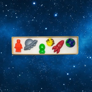 May include: A set of six space-themed crayons in a cardboard box. The crayons are shaped like an astronaut, a spaceship, a planet, a rocket, and a UFO. The crayons are red, blue, green, yellow, and gray.
