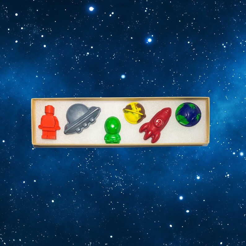 May include: A set of six space-themed crayons in a cardboard box. The crayons are shaped like an astronaut, a spaceship, a planet, a rocket, and a UFO. The crayons are red, blue, green, yellow, and gray.