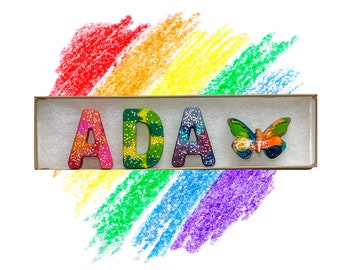 May include: A rainbow-colored crayon set with the letters "ADA" and a butterfly. The crayons are in a box with a white background.