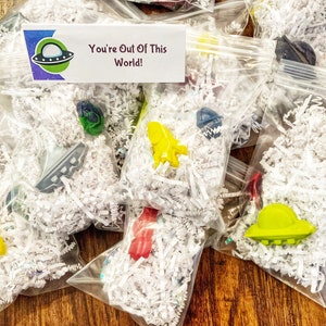 May include: A collection of small plastic bags filled with white shredded paper and colorful plastic toys. The bags have a tag that says "You're Out Of This World!" and a cartoon image of a UFO.