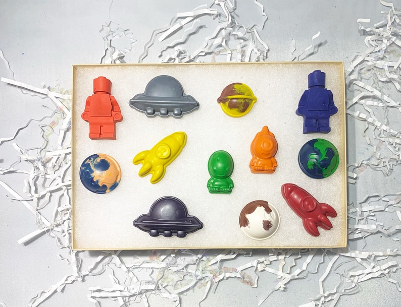 May include: A set of 12 space-themed crayons in a rectangular box. The crayons are shaped like planets, rockets, aliens, and spaceships. The crayons are in various colors, including red, blue, green, yellow, and orange.