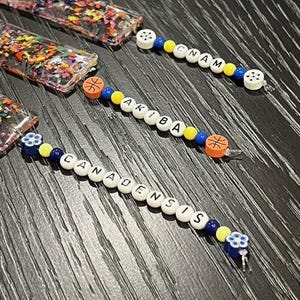 May include: Close-up of several beaded pacifier clips. The clips feature white letter beads spelling out names, along with colorful beads and decorative accents. The clips have a basketball and flower design.