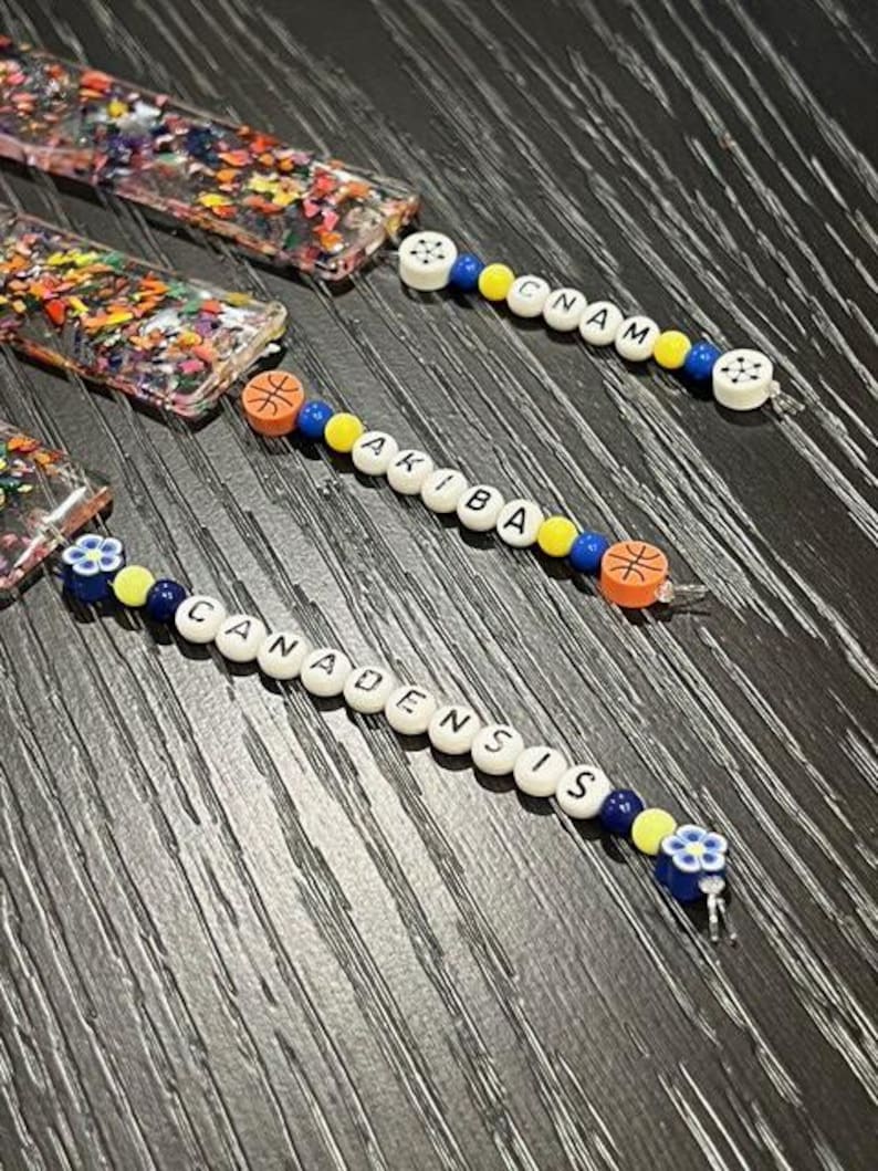 May include: Close-up of several beaded pacifier clips. The clips feature white letter beads spelling out names, along with colorful beads and decorative accents. The clips have a basketball and flower design.