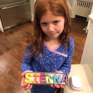 May include: A child holds a box with the name "SIENNA" spelled out in colourful, glitter-covered letters. The letters are in bright colours, including yellow, red, green, blue, and purple. The child is wearing a blue pajama set with a star pattern.