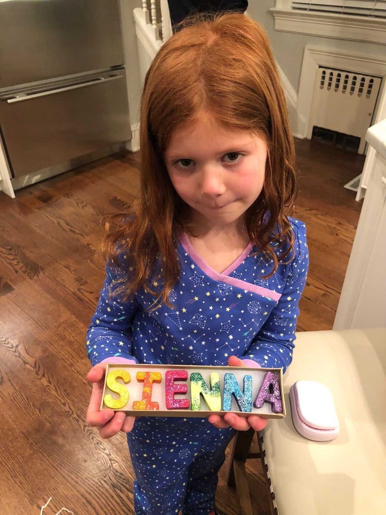 May include: A child holds a box with the name "SIENNA" spelled out in colourful, glitter-covered letters. The letters are in bright colours, including yellow, red, green, blue, and purple. The child is wearing a blue pajama set with a star pattern.