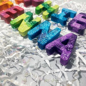 May include: A selection of colourful, glitter-covered letter-shaped objects in shades of red, yellow, green, blue, and purple. The letters are arranged on a bed of white shredded paper. The letters spell out the word "THANKS", creating a bright and appealing display.