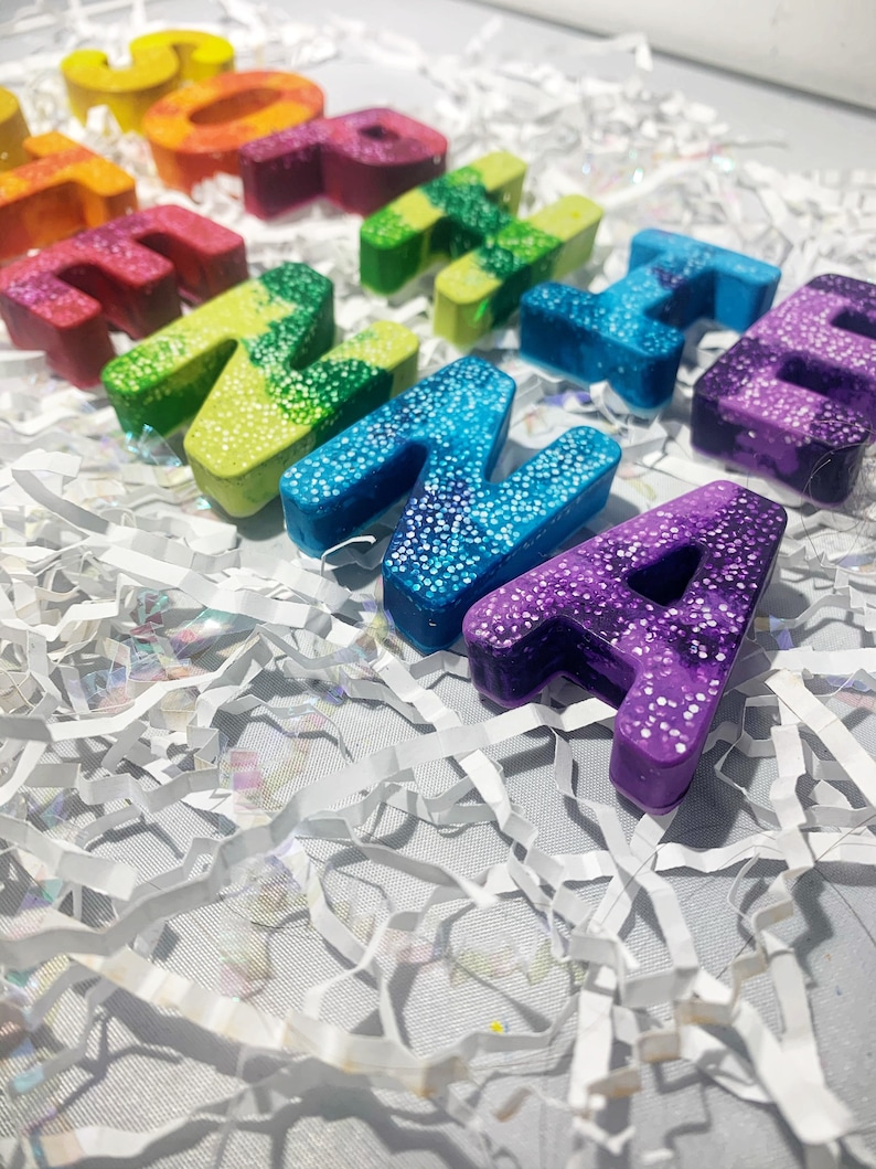 May include: A selection of colourful, glitter-covered letter-shaped objects in shades of red, yellow, green, blue, and purple. The letters are arranged on a bed of white shredded paper. The letters spell out the word "THANKS", creating a bright and appealing display.