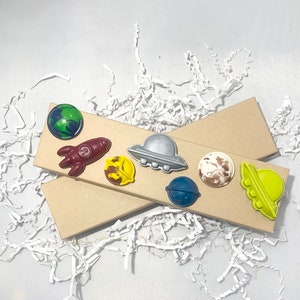 May include: A set of eight space-themed crayons in a brown cardboard box. The crayons are shaped like planets, rockets, and UFOs. The crayons are a variety of colors, including blue, green, yellow, red, and brown.