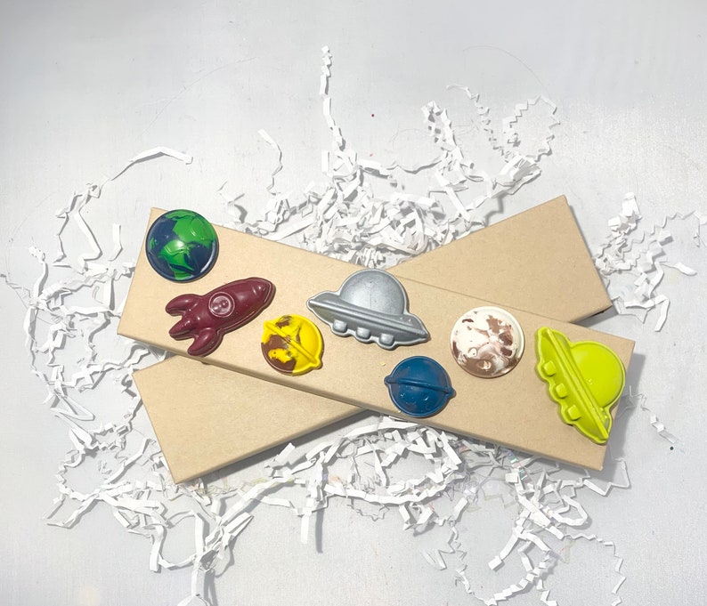 May include: A set of eight space-themed crayons in a brown cardboard box. The crayons are shaped like planets, rockets, and UFOs. The crayons are a variety of colors, including blue, green, yellow, red, and brown.