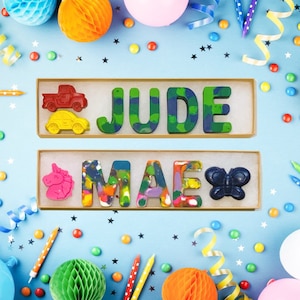 May include: Two wooden boxes with colorful crayon letters spelling out the names "JUDE" and "MAE". The boxes are on a blue background with confetti and other decorations.