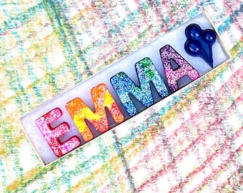 May include: A rectangular box contains the name "EMMA" spelled out in colorful, glitter-covered crayons. The crayons are in shades of pink, orange, blue, and green. A cluster of three blue crayon shapes is at the end of the box.