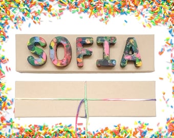 May include: A brown cardboard box with a colorful crayon name spelling "SOFIA" on top. The box is tied with a string and surrounded by colorful sprinkles.