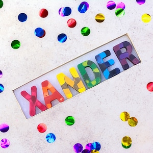 May include: A box of colorful crayons spelling the name "XANDER". Each letter is a different color, including red, orange, yellow, green, blue, and purple. The crayons are in a rectangular box, surrounded by colorful confetti.