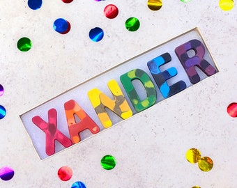 May include: A box of colorful crayons spelling the name "XANDER". Each letter is a different color, including red, orange, yellow, green, blue, and purple. The crayons are in a rectangular box, surrounded by colorful confetti.