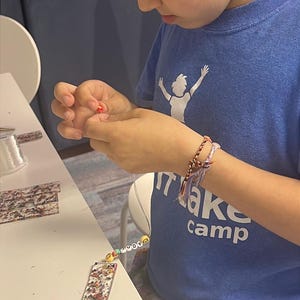 May include: A young person is crafting, holding a small red bead. They wear a blue t-shirt with "Make Camp" printed on it and a beaded bracelet. A decorated bookmark and beads are on the table.