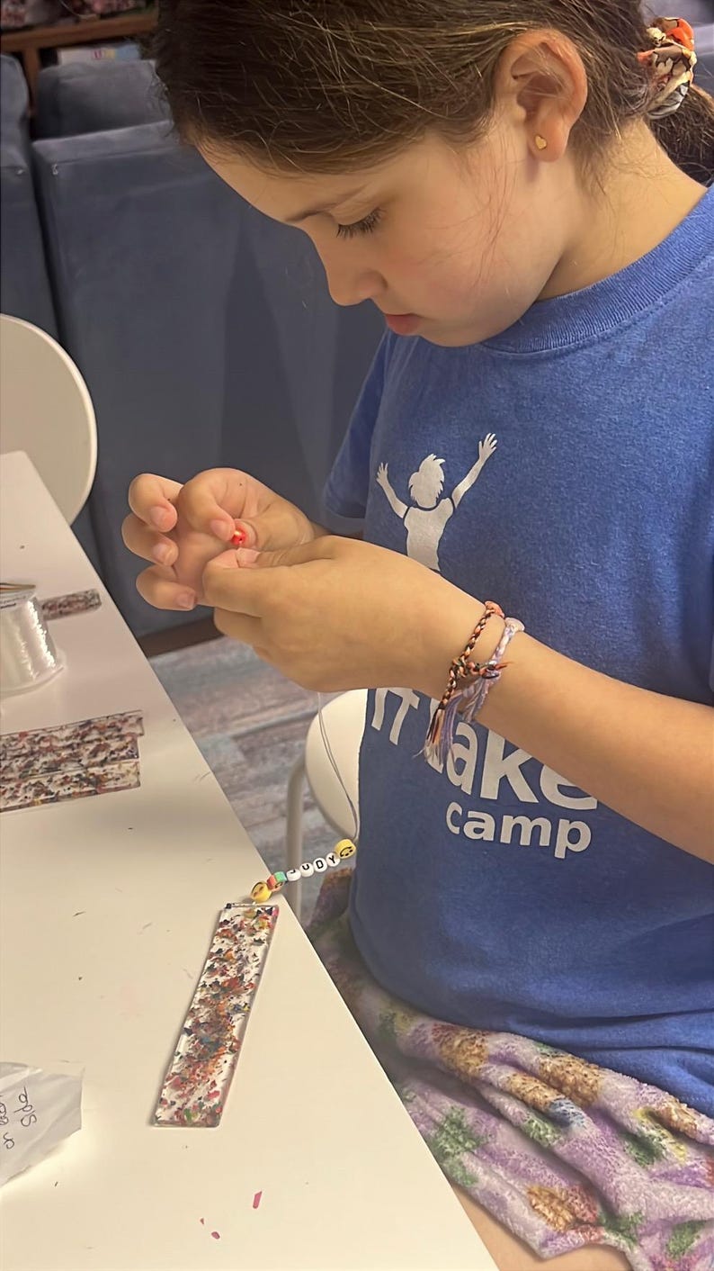May include: A young person is crafting, holding a small red bead. They wear a blue t-shirt with "Make Camp" printed on it and a beaded bracelet. A decorated bookmark and beads are on the table.