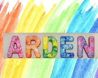 May include: A rectangular sign with the name "ARDEN" spelled out in colorful, glitter-covered letters. The letters are arranged horizontally against a rainbow-colored watercolor background. The letters are in shades of pink, orange, yellow, green, blue, and purple.