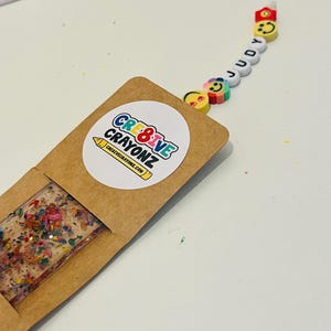 May include: A handmade bookmark with a rectangular resin design filled with colorful crayon shavings. The bookmark is in a brown paper sleeve with a round sticker that says "CRE8IVE CRAYONZ" and "creativecrayonz.com". A beaded charm spells "JUDY" with smiley face beads.