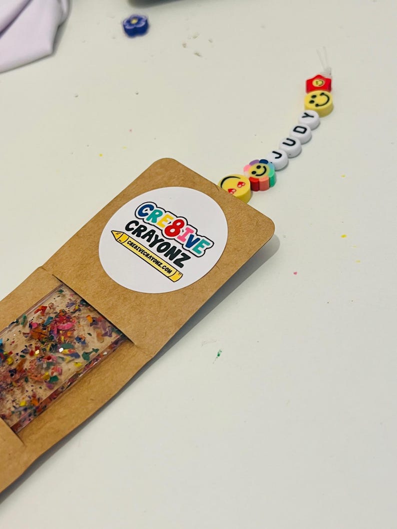 May include: A handmade bookmark with a rectangular resin design filled with colorful crayon shavings. The bookmark is in a brown paper sleeve with a round sticker that says "CRE8IVE CRAYONZ" and "creativecrayonz.com". A beaded charm spells "JUDY" with smiley face beads.