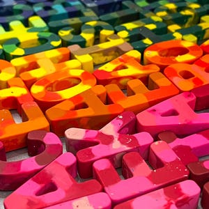 May include: A close-up of letter-shaped crayons in a rainbow of colours. The crayons are arranged in rows, with each letter a solid block of colour. The colours range from red and orange to green, blue, and purple, ideal for art projects.