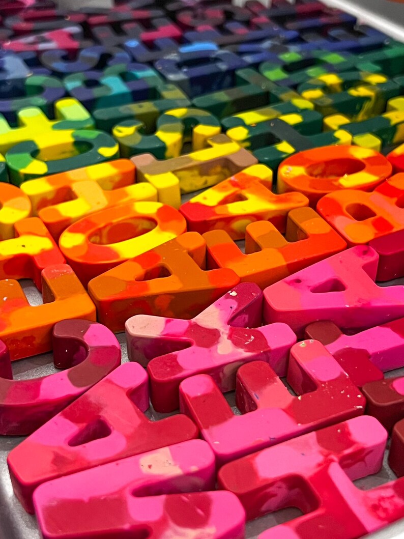 May include: A close-up of letter-shaped crayons in a rainbow of colours. The crayons are arranged in rows, with each letter a solid block of colour. The colours range from red and orange to green, blue, and purple, ideal for art projects.