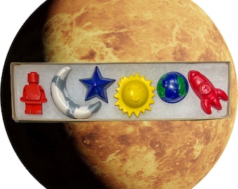 May include: A set of six colorful crayons shaped like a rocket, a star, a sun, a crescent moon, a globe, and a red Lego figure. The crayons are arranged in a white box with a brown lid.