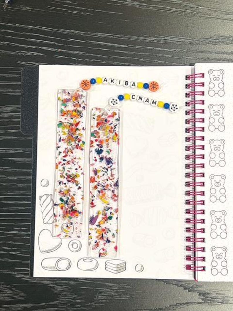 May include: Two clear resin bookmarks filled with colorful confetti sit on a white notebook page. The word "AKIBA CNAM" is spelled out with colorful beads. The notebook has a spiral binding and a pattern of bear illustrations.