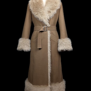 May include: A long, tan coat with a faux fur collar, cuffs, and hem. The coat features a matching belt with a silver buckle. The coat is displayed on a mannequin against a black background.